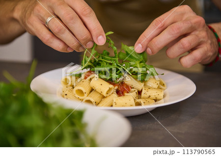 Close-up hands of a chef in home Kitchen, preparing and decorating meal with fresh arugula leaves, preparing pasta with tomato sauce according to traditional Italian recipe. Cuisine. Culinary. Epicure 113790565