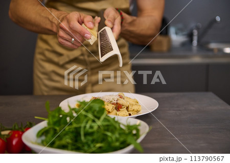 Closeup man cooking dinner at home, preparing Italian pasta with tomato sauce and pouring grated parmesan cheese. A bowl with fresh clean arugula leaves on the foreground 113790567