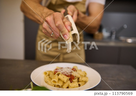Focus on hands of a male chef in an beige apron, grating parmesan cheese into a dish of spaghetti with tomato sauce, cooking dinner according to traditional Italian recipe in the home kitchen. Focus on hands of a male chef in an beige apron, grating parmesan cheese into a dish of spaghetti with tomato sauce, cooking dinner according to traditional Italian recipe in the home kitchen. 113790569