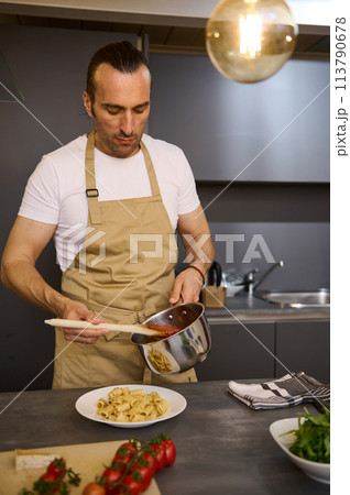 Vertical shot of attractive European male chef pouring tomato sauce on Italian pasta, plating up the dish before serving. Man cooking dinner at home kitchen. Cuisine. Culinary. Epicure. Food concept 113790678