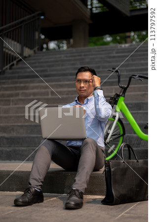 A determined Asian businessman sits on steps, working on a laptop, deep in thought. A determined Asian businessman sits on steps, working on a laptop, deep in thought. 113797592