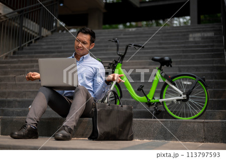 A confused Asian businessman sits on steps with a laptop, showing puzzled expression. A confused Asian businessman sits on steps with a laptop, showing puzzled expression. 113797593