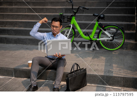 A determined Asian businessman sits on steps outside with his laptop, showing his fist, celebrating. A determined Asian businessman sits on steps outside with his laptop, showing his fist, celebrating. 113797595