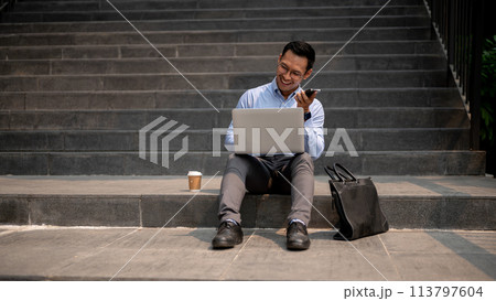 A businessman sits on steps, sending voice message via his smartphone while working on his laptop. A businessman sits on steps, sending voice message via his smartphone while working on his laptop. 113797604