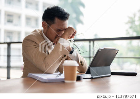 A frustrated Asian businessman covers his face with his hands while looking at a digital tablet. 113797671