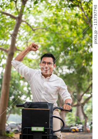 An excited Asian businessman rides a bike triumphantly, raising his fist in celebration. An excited Asian businessman rides a bike triumphantly, raising his fist in celebration. 113797748