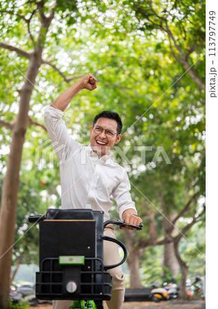 An excited Asian businessman rides a bike triumphantly, raising his fist in celebration. 113797749