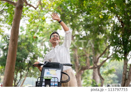 A happy businessman smiles and waves while pushing his bike, greeting someone while heading to work. A happy businessman smiles and waves while pushing his bike, greeting someone while heading to work. 113797755