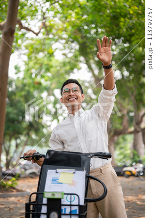 A happy businessman smiles and waves while pushing his bike, greeting someone while heading to work. A happy businessman smiles and waves while pushing his bike, greeting someone while heading to work. 113797757