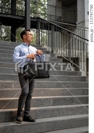 An exhausted Asian millennial businessman stands on the stairs, holding a coffee cup and a briefcase An exhausted Asian millennial businessman stands on the stairs, holding a coffee cup and a briefcase 113797790