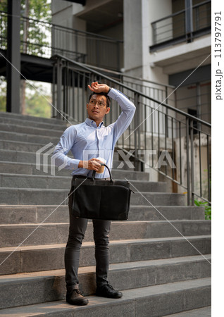 An exhausted Asian millennial businessman stands on the stairs, holding a coffee cup and a briefcase An exhausted Asian millennial businessman stands on the stairs, holding a coffee cup and a briefcase 113797791