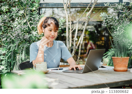 Contemplative woman works on her laptop at a wooden garden table, a serene green setting with plants and sunlight enhancing the calm work environment. 113800682