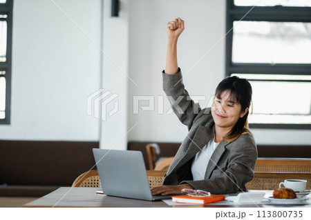 Businesswoman in office attire celebrates a success with a fist pump in front of her laptop at her desk. Businesswoman in office attire celebrates a success with a fist pump in front of her laptop at her desk. 113800735