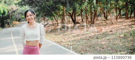 Young woman in a white crop top and pink leggings stands smiling on a sunlit park pathway with greenery in the background. 113800736