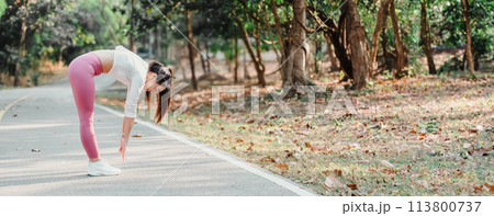 Woman in pink leggings touches her toes while stretching on a park path, surrounded by trees and fallen leaves. 113800737