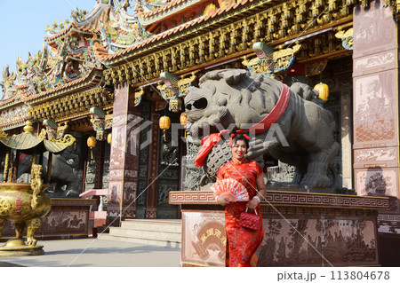 Portrait pretty Asian female in red traditional Chinese costume holding a fan standing and posing at stone lion  in the shrine. 113804678