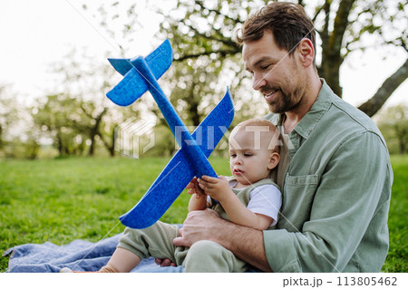 Father and toddler playing with foam glider plane. Single dad having fun with baby during warm spring day. Father's day concept. Father and toddler playing with foam glider plane. Single dad having fun with baby during warm spring day. Father's day concept. 113805462