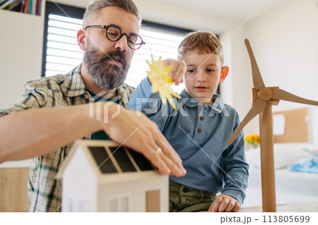 Father explaining renewable energy, teaching about sustainable lifestyle his young son. Playing with house with solar panels, wind turibine at home. 113805699