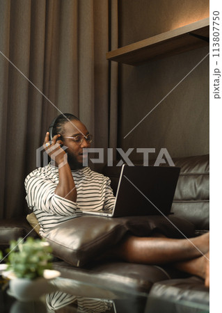 African man wearing headphone watching movie on laptop at home 113807750