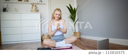 Portrait of young charismatic fitness trainer, girl blogger records video on digital camera, talks about health and workout, doing exercises on rubber mat in a room at home 113810375