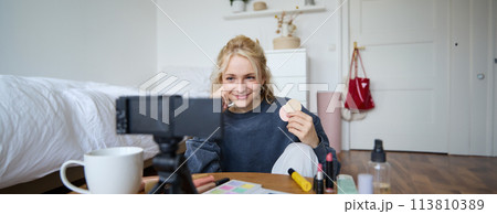 Image of young woman, vlogger records video on digital camera, shows beauty products, recommending makeup for audience on social media, sits on front in a room Image of young woman, vlogger records video on digital camera, shows beauty products, recommending makeup for audience on social media, sits on front in a room 113810389