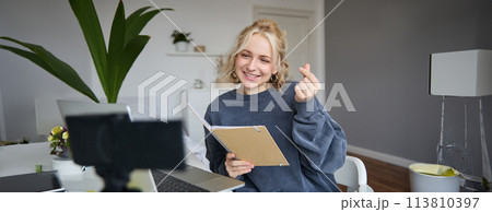 Cute young woman in her room, holds notebook, sits with laptop, looks at digital camera, records vlog or online tutorial 113810397