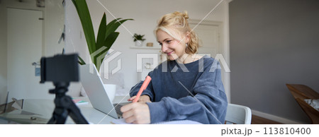 Portrait of smiling blond woman writing in notebook, making notes, recording content for social medial on digital camera, looking at laptop, working or studying Portrait of smiling blond woman writing in notebook, making notes, recording content for social medial on digital camera, looking at laptop, working or studying 113810400