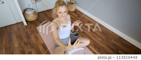 Portrait of young blogger, fitness instructor showing how to do exercises, sitting on yoga mat with smartphone, talking to followers on live stream 113810448