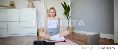 Portrait of young fitness instructor, vlogger showing exercises on camera, recording herself, sitting on mat with laptop, doing workout, explaining yoga movements to followers Portrait of young fitness instructor, vlogger showing exercises on camera, recording herself, sitting on mat with laptop, doing workout, explaining yoga movements to followers 113810475
