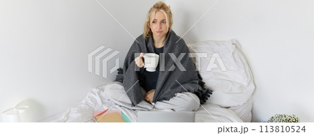 Portrait of woman catching a flu, sneezing, feeling sick, sitting on bed with laptop and working on remote while caught a cold Portrait of woman catching a flu, sneezing, feeling sick, sitting on bed with laptop and working on remote while caught a cold 113810524