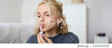 Close up portrait of beautiful blond woman applies lip gloss on her lips, puts on makeup, gets ready for night out, using beauty products, looks in mirror 113810530