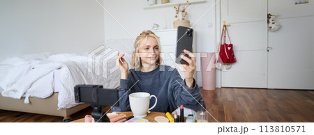 Portrait of young beauty blogger, recording video on digital camera, doing online live stream while applying makeup, talking to her followers, sitting on bedroom floor 113810571