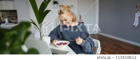 Portrait of smiling blond young woman, eating in front of laptop, watching videos online while having breakfast, enjoying dessert, sitting in bedroom Portrait of smiling blond young woman, eating in front of laptop, watching videos online while having breakfast, enjoying dessert, sitting in bedroom 113810612