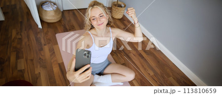 Portrait of young sporty woman taking selfie during workout training, content creator takes photos, shows muscles on camera, flexing biceps 113810659