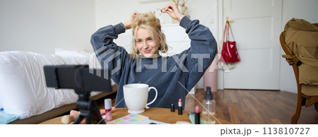 Lifestyle blogger records video of herself while doing hairstyle, showing makeup lifehacks for social media followers, vlogging, using camera for her blog, sitting in a room, drinking tea Lifestyle blogger records video of herself while doing hairstyle, showing makeup lifehacks for social media followers, vlogging, using camera for her blog, sitting in a room, drinking tea 113810727
