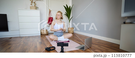 Portrait of young fitness blogger, woman showing exercises to her followers, recording video on digital camera, doing workout training session, home yoga, sitting in front of laptop on rubber mat 113810777