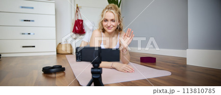 Portrait of smiling fitness instructor, social media content creator recording a vlog about sport, using rubber yoga mat, waving hand and looking at digital video camera Portrait of smiling fitness instructor, social media content creator recording a vlog about sport, using rubber yoga mat, waving hand and looking at digital video camera 113810785
