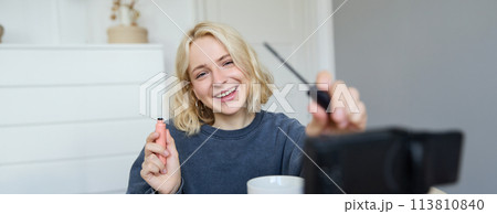 Image of cheerful, beautiful young lifestyle blogger, woman sitting on floor and recording video about makeup, holding mascara, making lifestyle content for her social media account and followers 113810840
