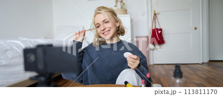 Portrait of smiling young woman applies makeup, shows beauty tutorial on video, records vlog on digital camera in her room, recommends cosmetic products 113811170