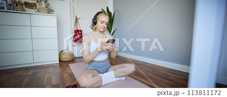 Portrait of young blond woman in headphones, turning on yoga, workout app on smartphone, choosing music on mobile phone application, sitting on rubber mat in room Portrait of young blond woman in headphones, turning on yoga, workout app on smartphone, choosing music on mobile phone application, sitting on rubber mat in room 113811172