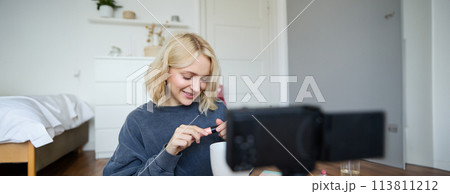 Close up portrait of smiling young woman with video camera in her room, showing mascara, reviewing beauty products for social media account. Beauty blogger creates vlog about makeup 113811212