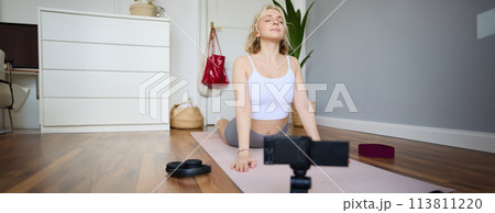 Portrait of young blogger, yoga content creator, showing exercises, recording video of herself working out at home on rubber mat 113811220