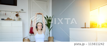 Portrait of young fitness woman doing warm-up exercises, looking at workout video on laptop, stretching arms, wearing headphones 113811242