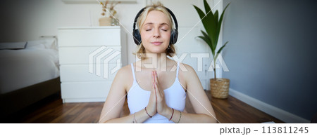 Portrait of woman meditating at home, sitting in headphones, listening to yoga podcast, holding hands in namaste sign Portrait of woman meditating at home, sitting in headphones, listening to yoga podcast, holding hands in namaste sign 113811245