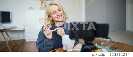 Smiling young beautiful lifestyle blogger, girl records a beauty vlog in her room, using professional video camera, showing makeup cosmetics to her followers, creates content for social media 113811284