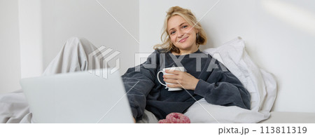 Portrait of happy woman enjoying day-off at home, lying in bed, watching tv show on laptop, looking at screen and smiling, drinking tea and eating in bedroom Portrait of happy woman enjoying day-off at home, lying in bed, watching tv show on laptop, looking at screen and smiling, drinking tea and eating in bedroom 113811319