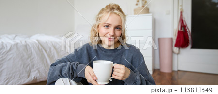 Portrait of young woman sitting on bedroom floor, drinking tea, holding white mug and smiling at camera Portrait of young woman sitting on bedroom floor, drinking tea, holding white mug and smiling at camera 113811349