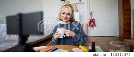 Portrait of cute blond woman, video content maker, vlogger recording on digital camera in bedroom, drinking cup of tea and talking to audience 113811472