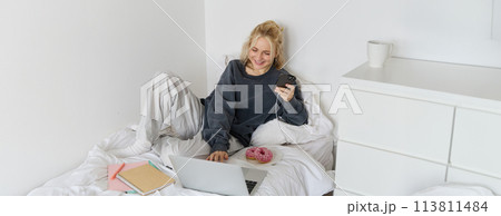 Portrait of smiling candid woman, lying in bed with doughnut, using smartphone and laptop, resting at home in bedroom, watching tv show or chatting online Portrait of smiling candid woman, lying in bed with doughnut, using smartphone and laptop, resting at home in bedroom, watching tv show or chatting online 113811484