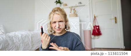 Portrait of young blogger, woman recording video about beauty, makeup products, showing lipstick and grimacing, express negative opinion, creating a lifestyle vlog content Portrait of young blogger, woman recording video about beauty, makeup products, showing lipstick and grimacing, express negative opinion, creating a lifestyle vlog content 113811504